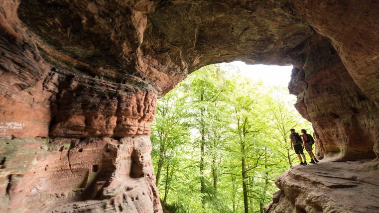 Zwei Wanderer stehen am Eingang der Genovevahöhle mit Blick auf grüne Bäume.