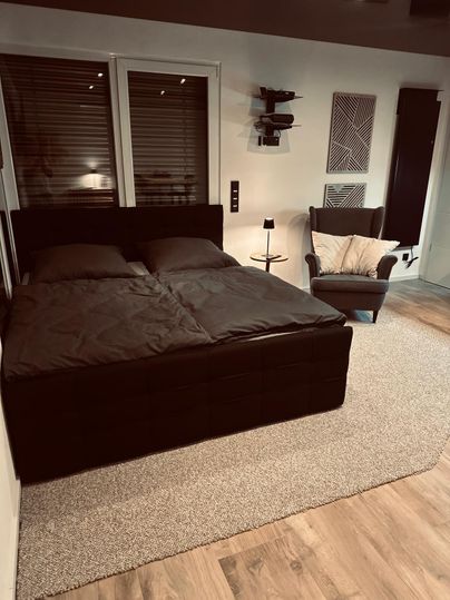 Ein modernes Schlafzimmer mit einem großen Doppelbett und einem gemütlichen Sessel. Der Boden ist mit einem Teppich bedeckt und es gibt Fenster mit Jalousien.