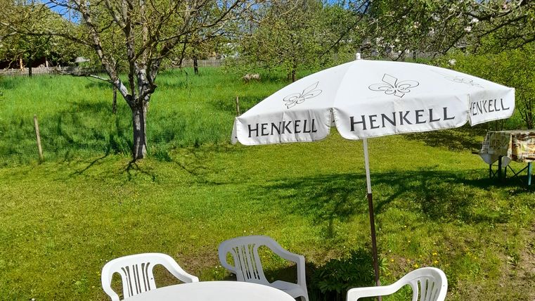 Ein gemütlicher Außenbereich mit einem runden Tisch und weißen Plastikstühlen. Ein großer Sonnenschirm bietet Schatten auf einer grünen Wiese.