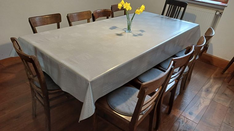 Ein großer Tisch mit mehreren Holzstühlen, der mit einer grauen Tischdecke bedeckt ist. In der Mitte steht eine Vase mit gelben Narzissen.