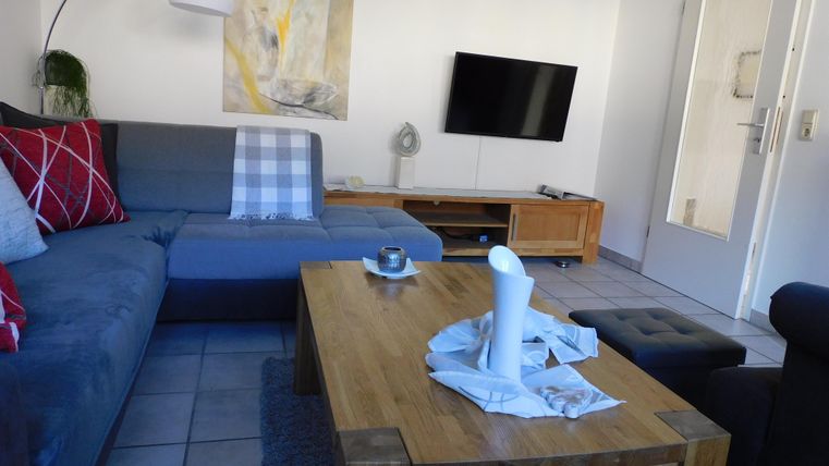 Ein gemütliches Wohnzimmer mit einem grauen Sofa und einem Holztisch. Im Hintergrund steht ein Fernseher und es gibt eine ansprechende Wanddekoration.