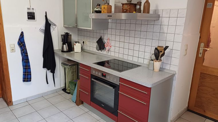 Eine moderne Küche mit roten Schränken und einer hellen Arbeitsplatte. Es gibt eine Kaffeemaschine, Kochutensilien und eine offene Tür im Hintergrund.