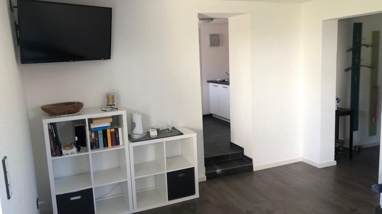 Ein heller Raum mit einem Fernseher an der Wand und einem Bücherregal. Im Hintergrund ist eine offene Tür zur Küche zu sehen.