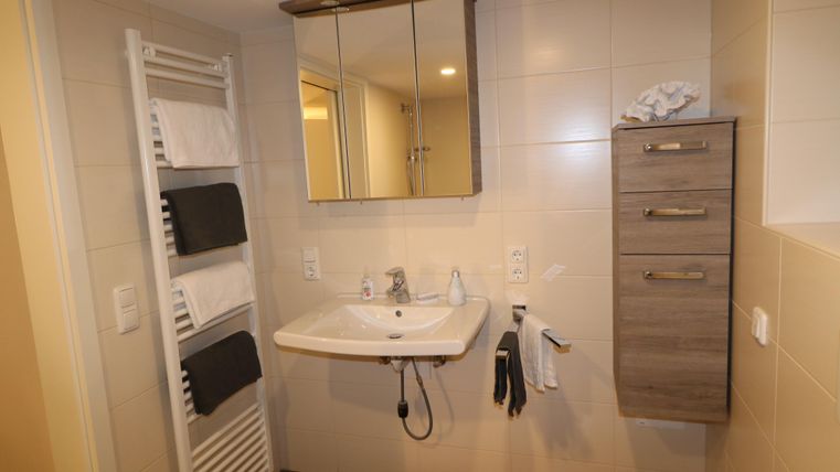 Ein modernes Badezimmer mit einem Waschbecken, einem Spiegel und einem Handtuchhalter. Die Wände sind hell und es gibt einen kleinen Holzschrank für Aufbewahrung.