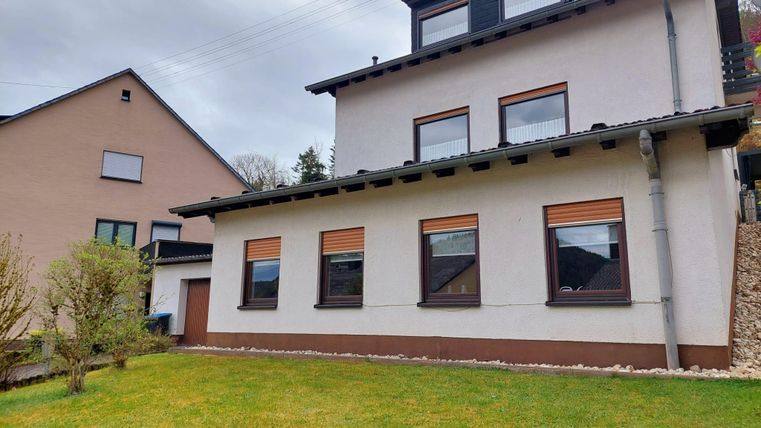 Ein modernes Haus mit mehreren Fenstern und einem Garten im Vordergrund. Es gibt auch ein benachbartes, traditionelles Gebäude.