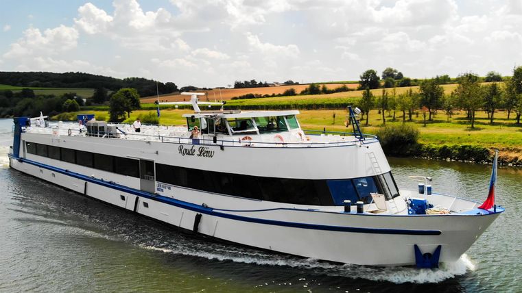 Ein weißes Schiff fährt auf einem ruhigen Fluss. Im Hintergrund sind grüne Felder und Bäume zu sehen.