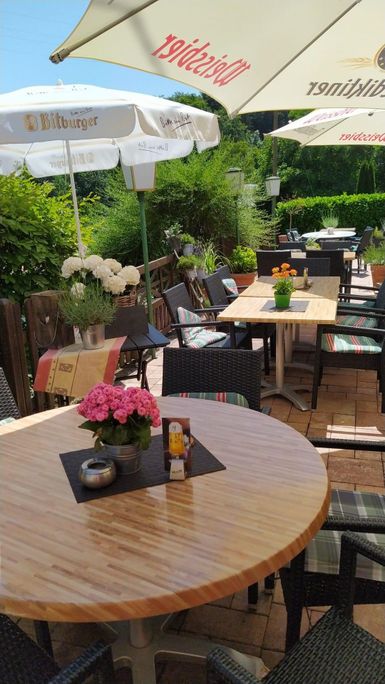 Ein einladender Außenbereich mit Tischen und Stühlen, umgeben von grünen Pflanzen. Auf den Tischen stehen Blumen und Sonnenschirme spenden Schatten.