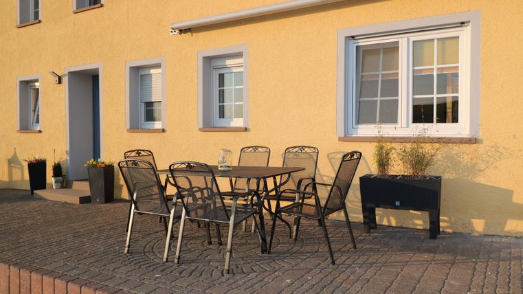 Ein gemütlicher Terrasse mit einem Tisch und sechs Stühlen. Die Wände des Gebäudes sind in einem warmen Gelbton gestrichen.