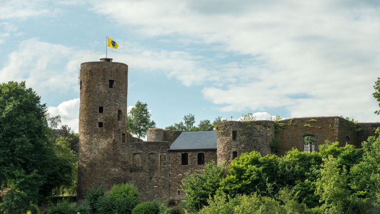 Burg Reuland mit Flagge auf dem Turm, umgeben von Bäumen.