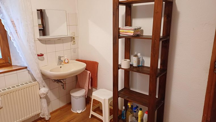 Ein einfaches Badezimmer mit einem Waschbecken und einem Spiegel. An der Wand steht ein Holzregal, das mit verschiedenen Pflegeprodukten gefüllt ist.