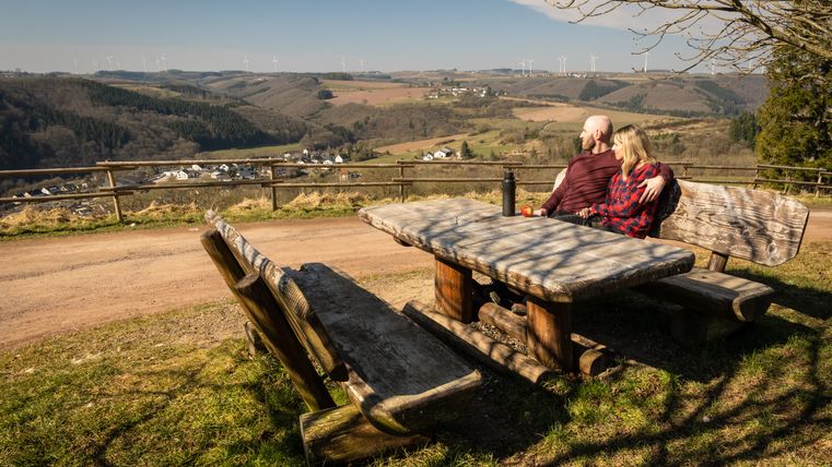Ein Paar sitzt auf einer Bank an einem Holztisch mit Blick auf eine hügelige Landschaft und Windräder in der Ferne.