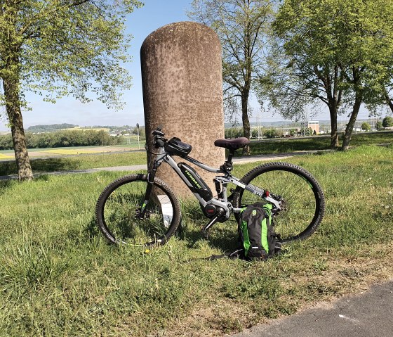 Un vélo est adossé à une colonne de pierre dans une prairie verdoyante. En arrière-plan, on aperçoit des arbres et un vaste paysage., © TI Bitburger Land, Steffi Wagner