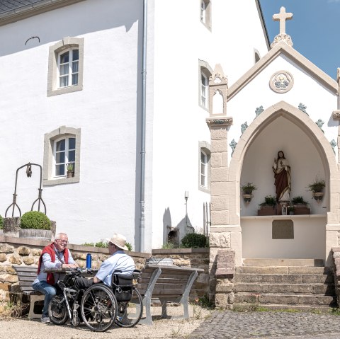 Two men, one in a wheelchair, sit on a bench in front of a white building with a religious statue in a niche. Sunny day., © Naturpark Südeifel, Thomas Urbany