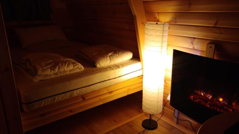 Ein gemütliches Schlafzimmer aus Holz mit einem Bett, zwei Kissen und einer modernen Stehlampe. Im Hintergrund befindet sich ein Elektrokamin, der eine warme Atmosphäre schafft.