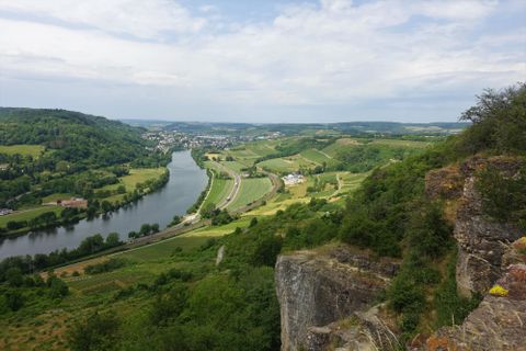 Eine malerische Landschaft mit einem Fluss, umgeben von grünen Hügeln und Weinbergen. Der Himmel ist bewölkt, was der Szene eine ruhige Atmosphäre verleiht.