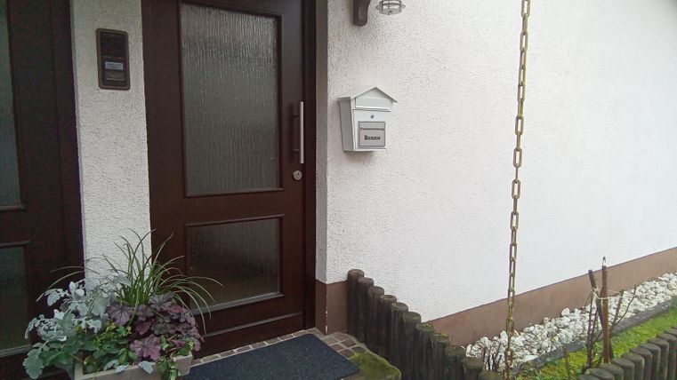 Der Eingangsbereich eines Hauses mit einer braunen Tür. Neben der Tür steht ein Pflanzenkübel und eine kleine Blumenrabatte ist sichtbar.