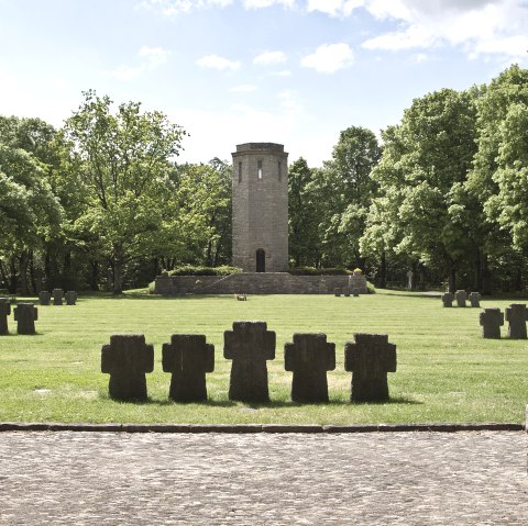 A war memorial with stone crosses on a meadow, a tower in the background, surrounded by green trees under a blue sky., © TI Bitburger Land