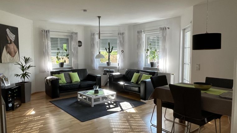 Ein modernes Wohnzimmer mit schwarzen Sofas und einem Esstisch. Große Fenster lassen viel Licht hinein und Pflanzen sorgen für eine frische Atmosphäre.