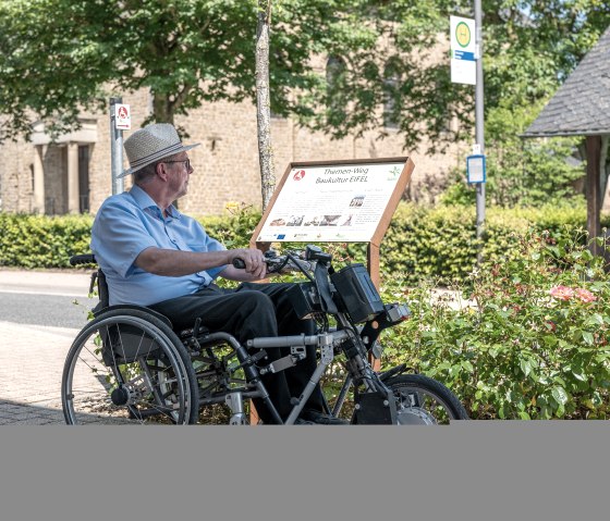 Een man in een rolstoel leest een bord voor het Eifel bouwcultuur themapad in Wolsfeld. Op de achtergrond zijn bomen en een gebouw te zien., &copy; Naturpark S&uuml;deifel, Thomas Urbany