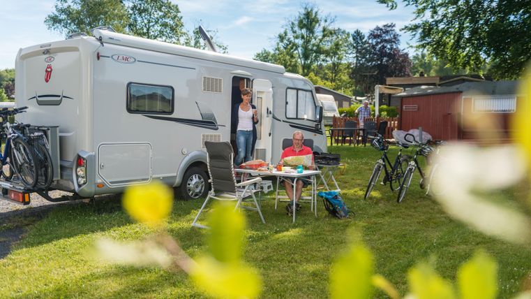 Ein Wohnmobil steht auf einem grünen Campingplatz. Menschen sitzen an einem Tisch im Freien und genießen ihre Zeit.