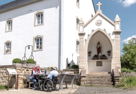 Zwei M&auml;nner, einer im Rollstuhl, sitzen auf einer Bank vor einem wei&szlig;en Geb&auml;ude mit einer religi&ouml;sen Statue in einer Nische. Sonniger Tag., &copy; Naturpark S&uuml;deifel, Thomas Urbany