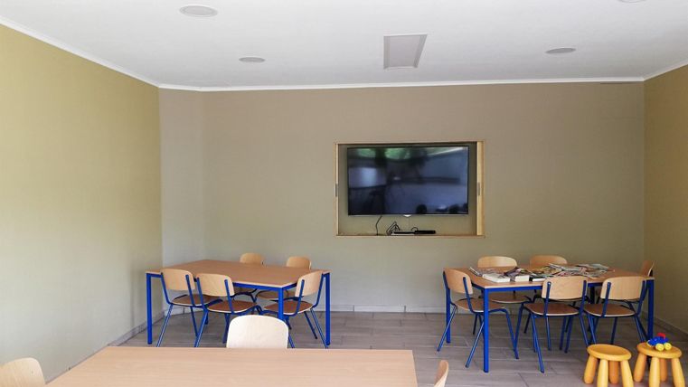 Ein heller Raum mit grauen Wänden und Holzfußboden. Es gibt einen Tisch mit Stühlen und einen Fernseher an der Wand.